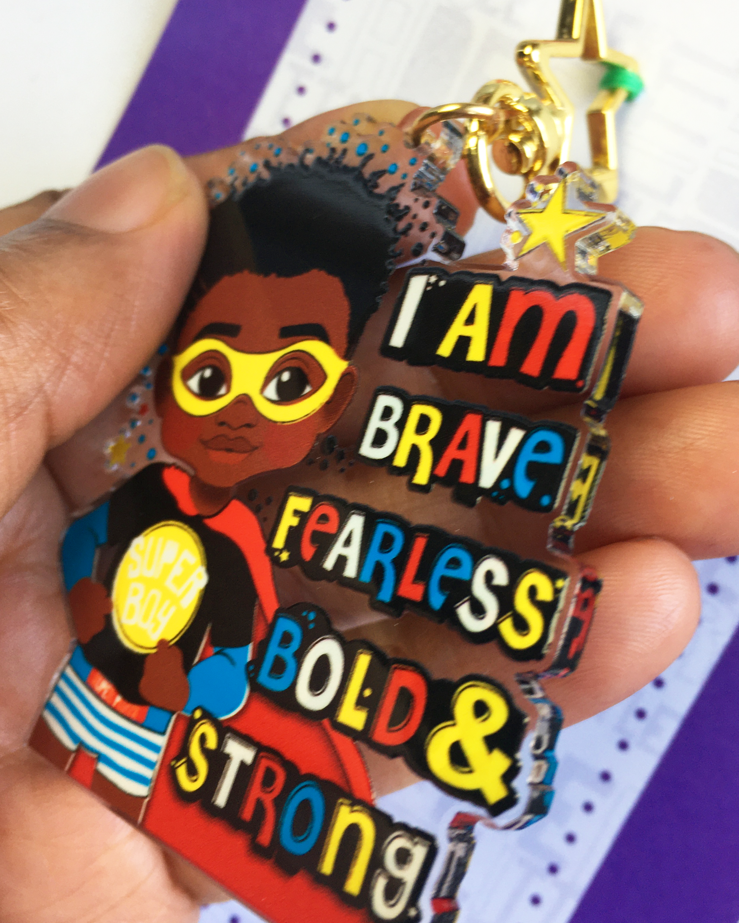 sideview of a black boy superhero keychain with motivational text held by a hand against a colourful background