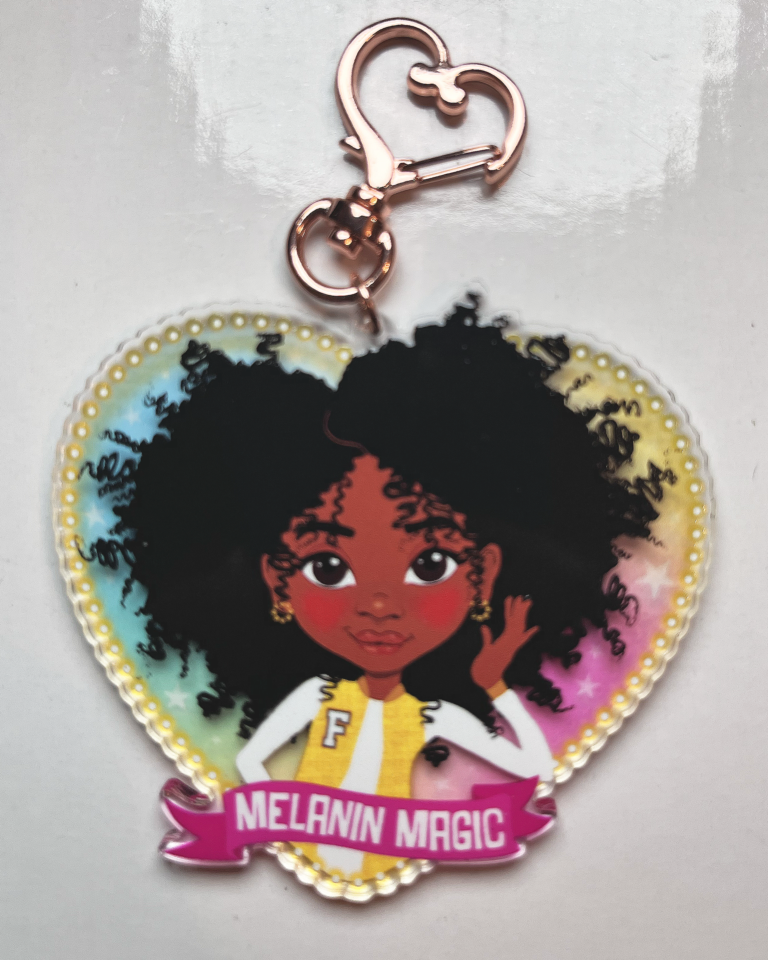 Heart-shaped keychain with a cartoon character and 'Melanin Magic' text on a transparent rainbow glazed background