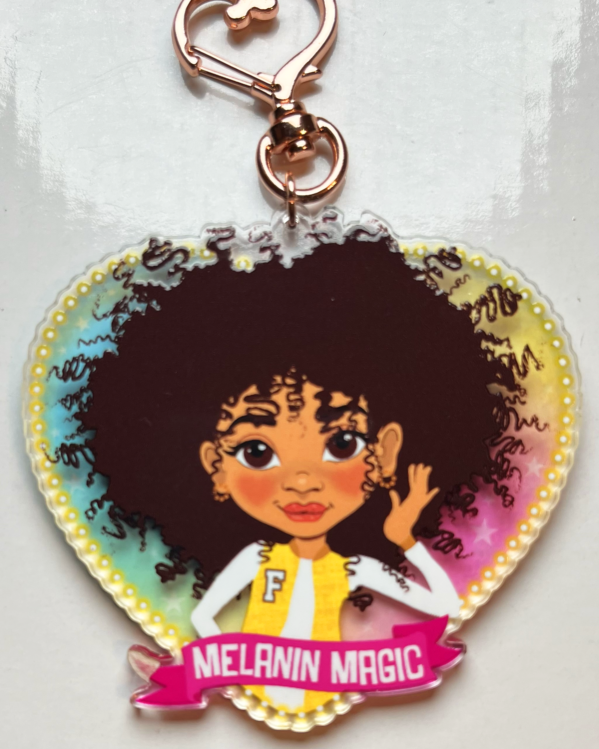 Heart-shaped keychain with a cartoon character and 'Melanin Magic' text on a transparent rainbow glazed background