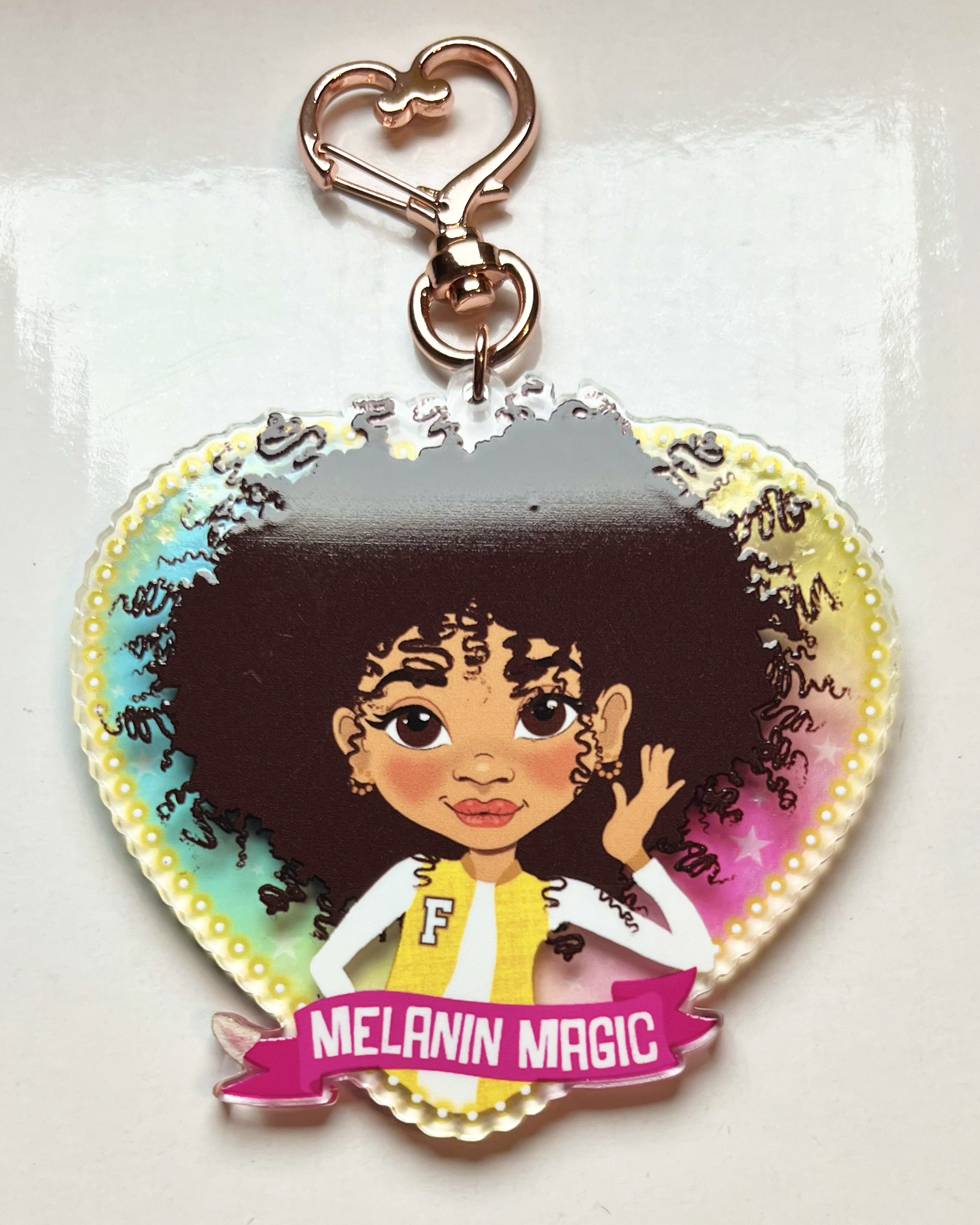 Heart-shaped keychain with a mixed race afro girl cartoon character and 'Melanin Magic' text on a transparent rainbow background