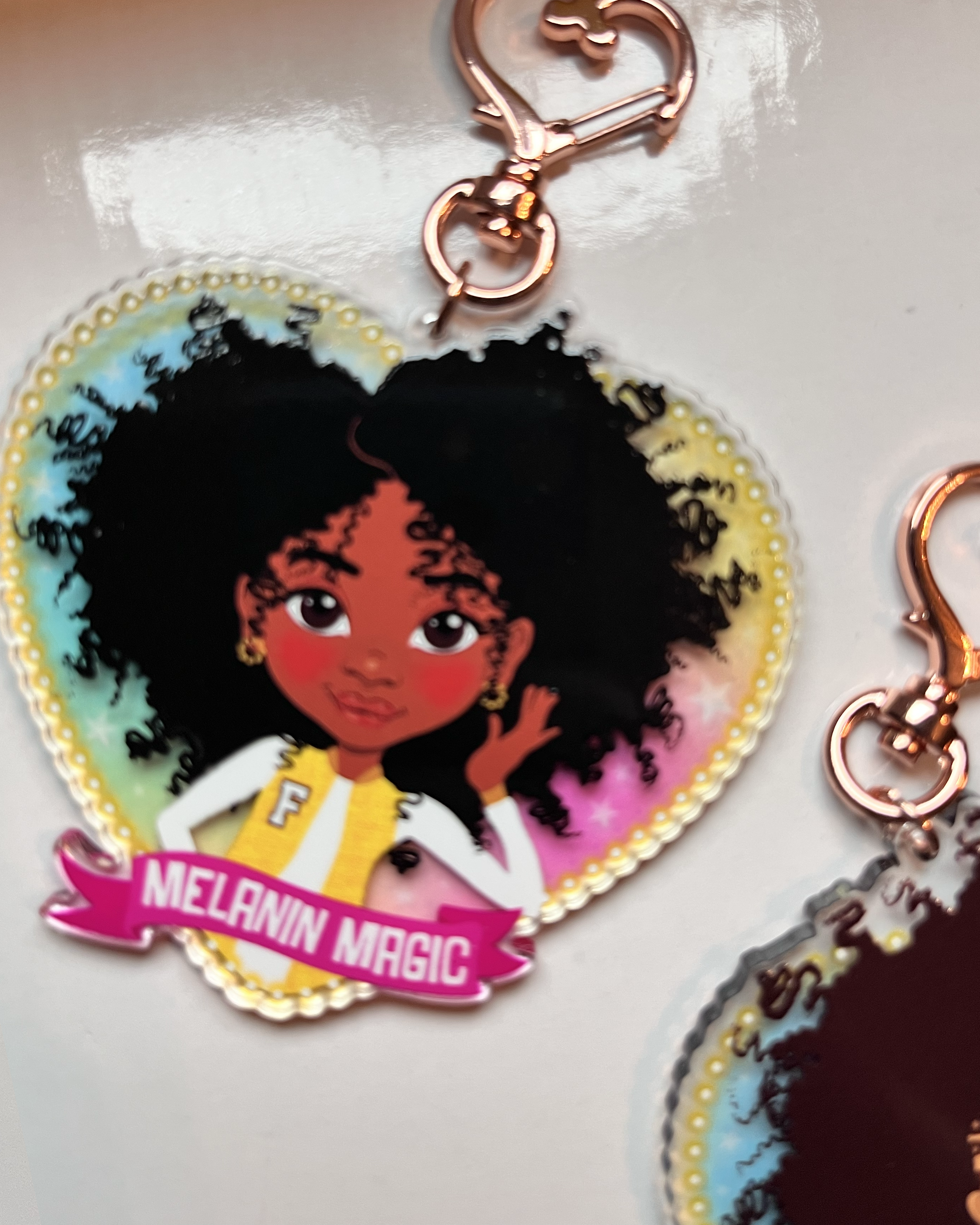 Heart-shaped keychain with a black afro girl cartoon character and 'Melanin Magic' text on a transparent rainbow background